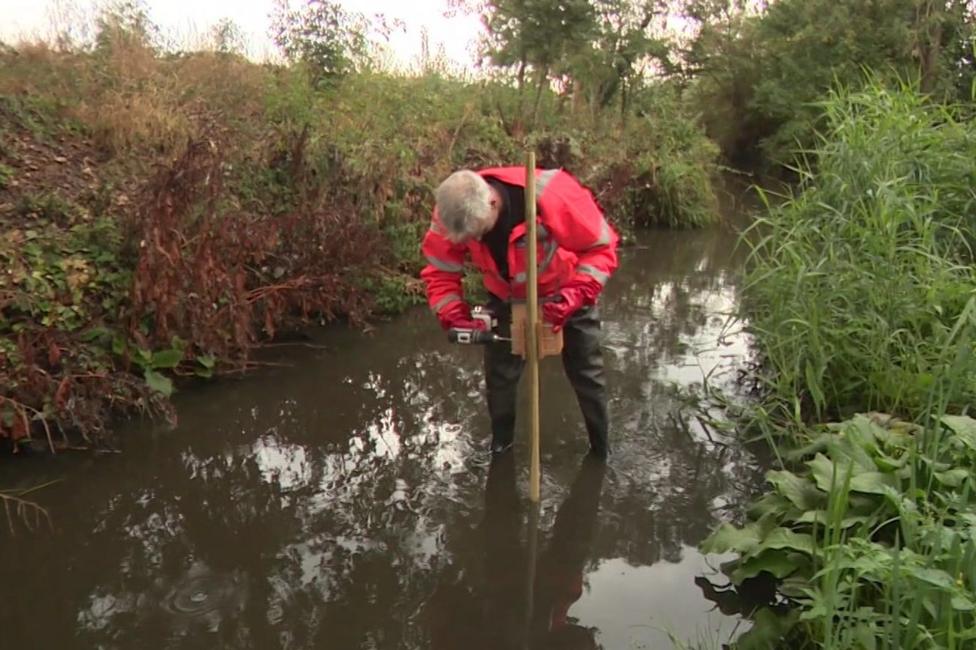 Man in a pink hi-viz jacket puts a stick into a murky stream