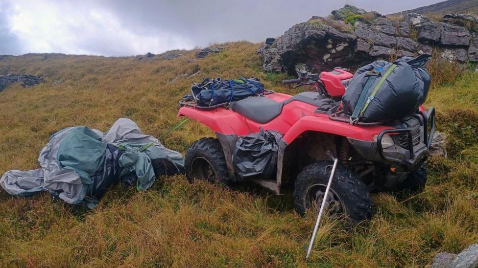 A red quadbike piled high with rubbish bags and discarded camping equipment in the middle of a grassy area.