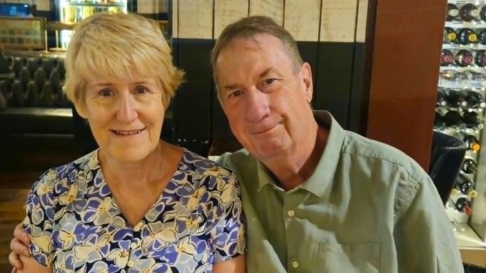 A man and woman sat next to each other in a restaurant. She has blonde hair and a floral top on. He is wearing a green shirt. There is a wine rack behind them.