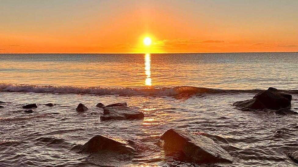 An orange sunrise over the water. There are rocks along the shore.