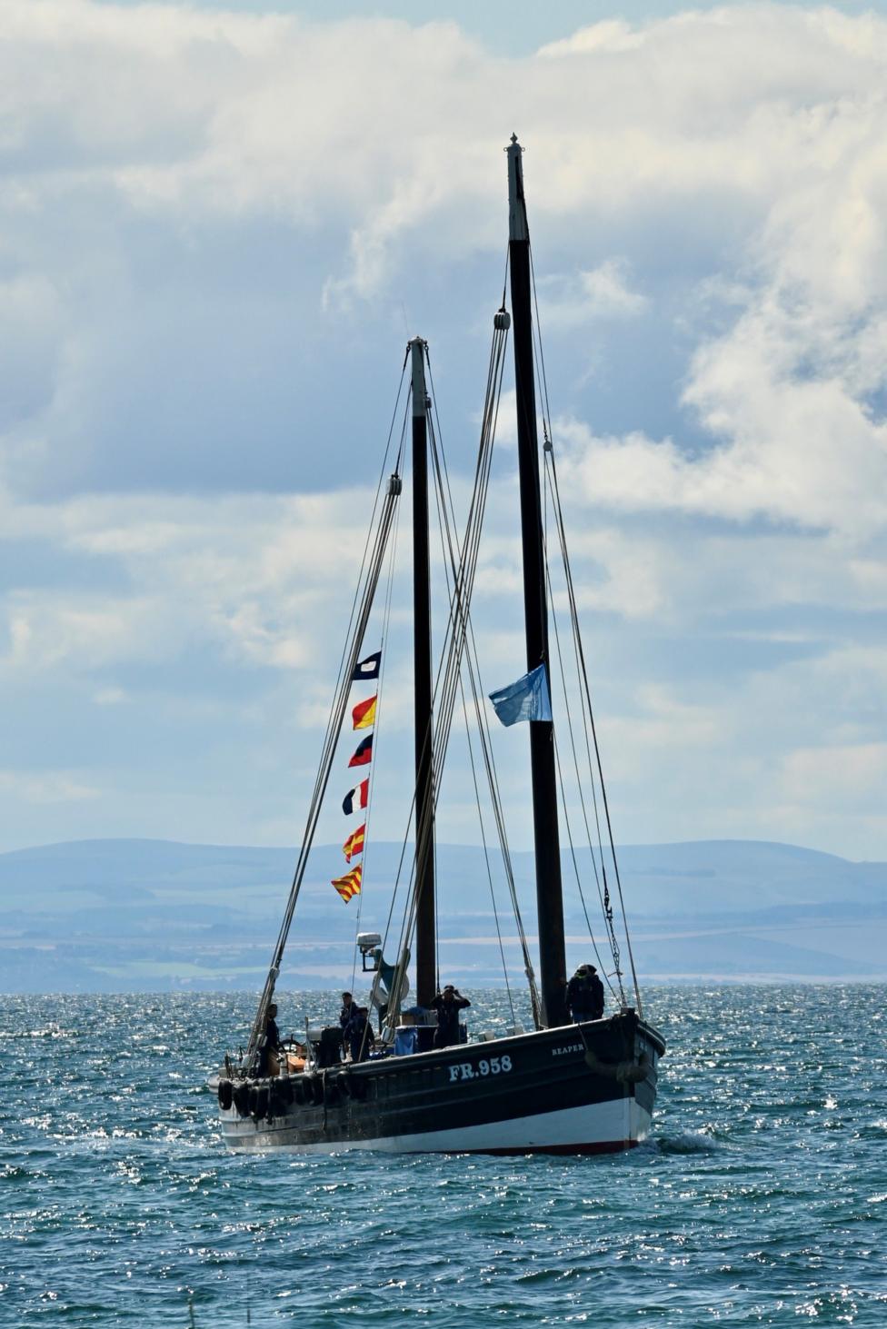 A 20th Century fishing boat in the water. It has two masts and bunting. The water has some waves and there are clouds in the sky.