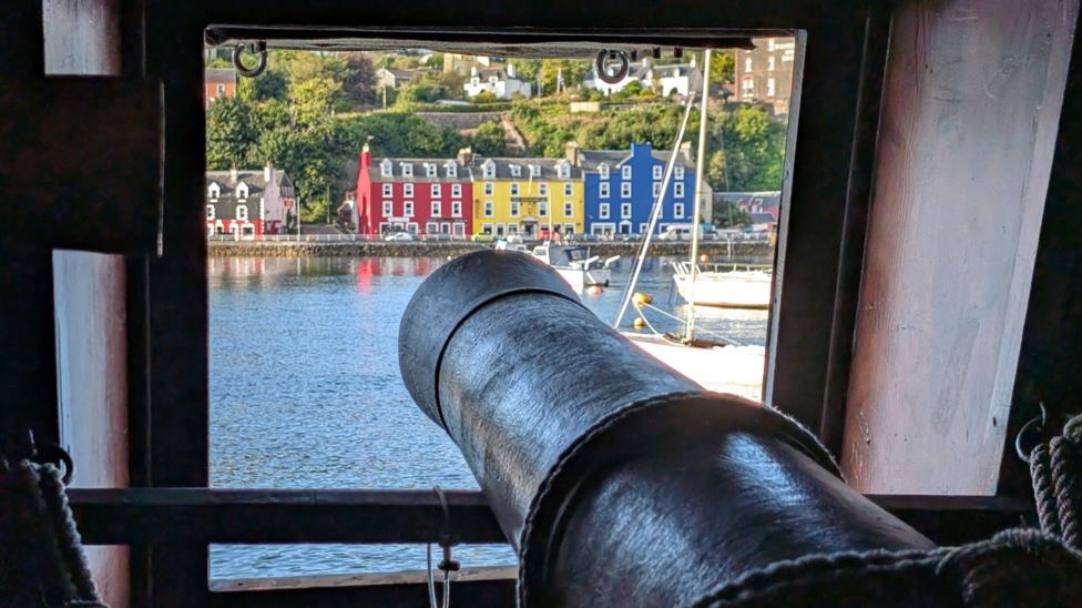 The view out of a window of the Galeon Andalucia. Bright coloured houses are on the shore front and there are boats in the water. A cannon is pointing out of the window.