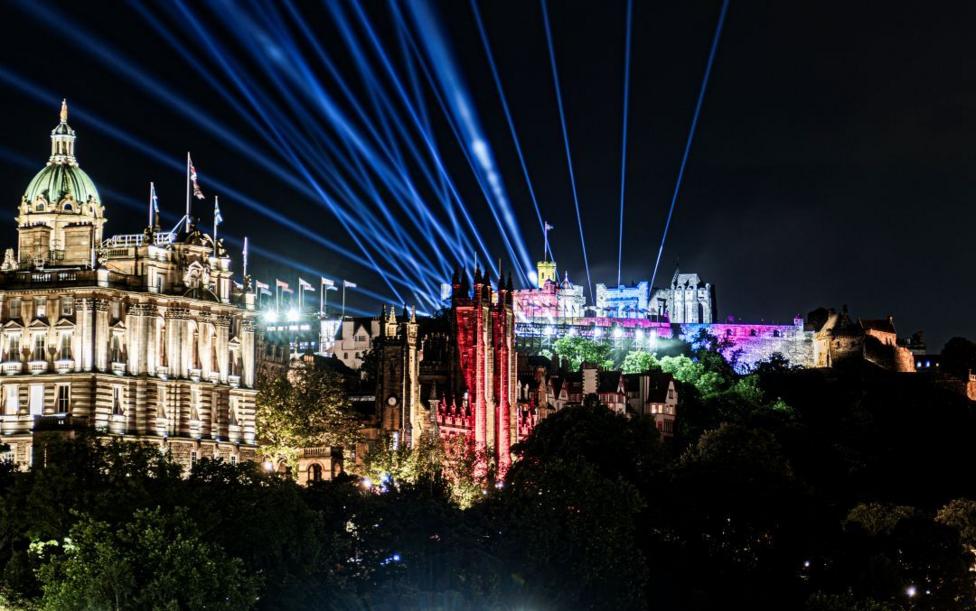 Edinburgh Castle lit up with different colours. Blue strobes point into the dark night sky. Other buildings in the foreground are lit up.