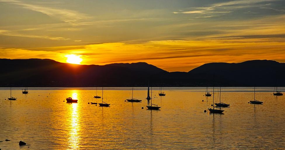 An orange sunset over the hills and reflected over the water. Silhouettes of boats are in the foreground.