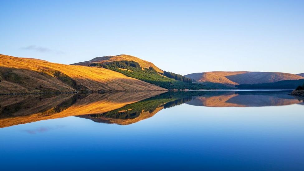 A reflection of some hills in clear water. There are no clouds in the sky of waves in the water. Trees line the hillside.