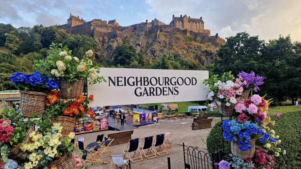 Neighbourhood Gardens in Edinburgh. The sign is surrounded by pots of bright coloured flowers and Edinburgh Castle stands in the background. There are deck chairs and tables inside.