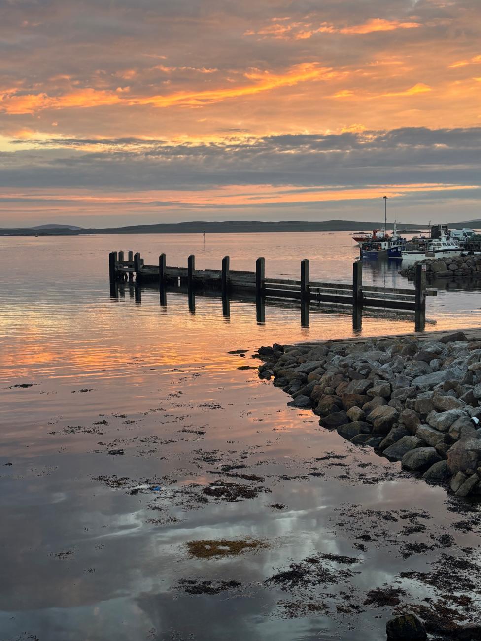 A pier with boats in the background and rocks in the foreground. The sky is cloudy with orange streaks, which are reflected on the calm water.