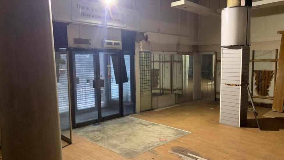 The ground floor of a vacant shop. the exit door is in the centre, with wooden floors and pillars close by