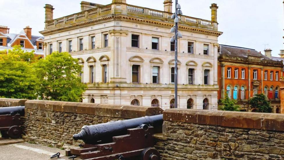 The former Danske Bank building is a large grey building. It is viewed from Derry's historic walls were cannons are seen up against the city walls. Copyright