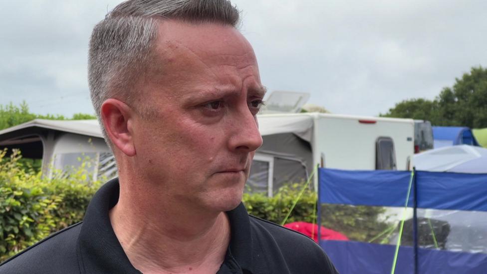 This is a close up of Stephen Brown. He is looking concerned. He has short, neat, grey hair and is wearing a black polo shirt. He is standing at the holiday park and there are caravans and tents behind him.