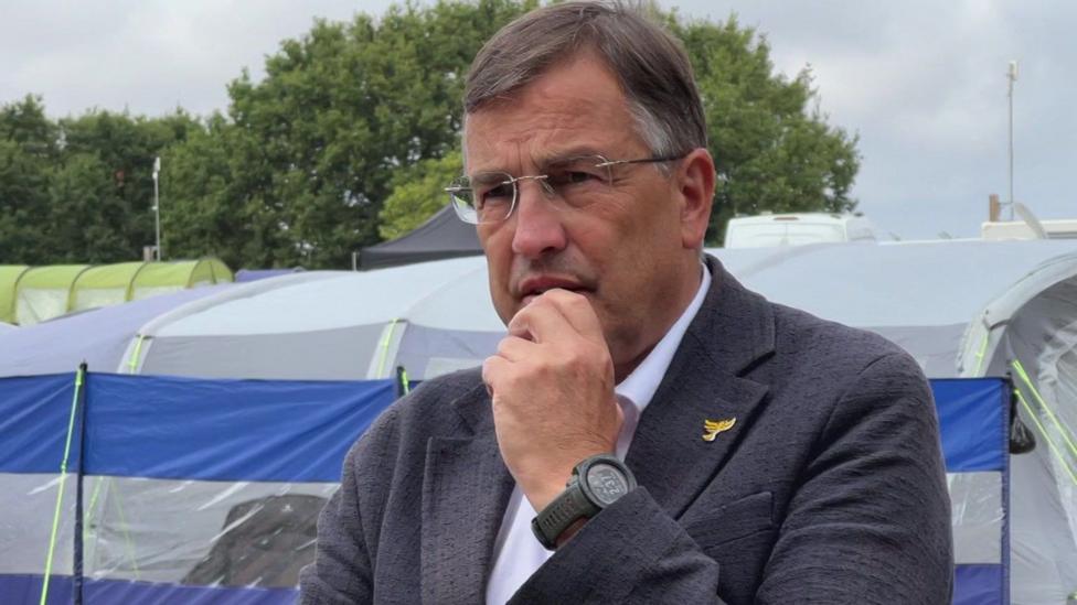Martin Wrigley is standing at the holiday park. He has short grey hair and rimless spectacles. He is wearing a grey blazer over a white shirt. He is holding his hand up to his mouth. There are rows of tents behind him and trees beyond them.