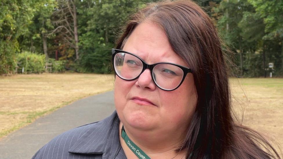 This is a close up of Rosie Dawson's face. She has long, dark brown hair and black spectacles. She is wearing a grey shirt with fine white stripes on it, and a green Devon County Council lanyard. She is standing on a path at the holiday park and there are trees in the distance.
