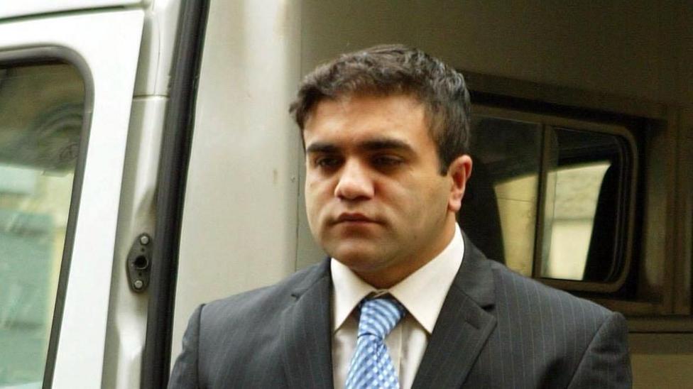 Imran Shahid is coming out of a prison vehicle. He has short, dark hair and is wearing a grey suit jacket, white shirt and blue and white checked tie.