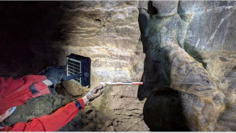 An archaeologist wearing a helmet, red boiler suit and black gloves, measures a ceiling artwork within a cave in south-west Ireland