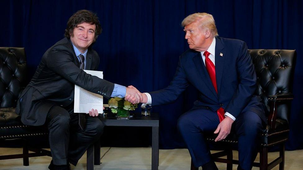 US President Donald Trump (R) shakes hands with President of Argentina Javier Milei during a bilateral meeting at the 80th session of the UN’s General Assembly at the United Nations headquarters on September 23, 2025 in New York City. The men are both wearing dark suits and sitting on chairs. Milei is smiling with his mouth closed, while Trump, looking at him, has a serious expression.