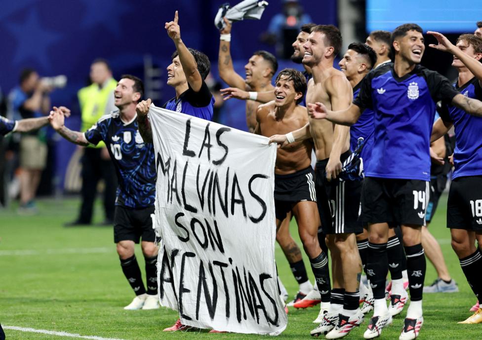 Football players, some with their shirts off, stand or jump while smiling on a green football pitch. Two of them hold a white banner that says in black writing "Las Malvinas Son Argentinas" - the Falklands are Argentina's.