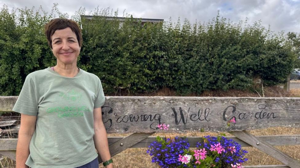 Dr Susan Taheri stood off the left of the centre looking and smiling at the camera, wearing a green t-shirt which says growing well garden. There is a fence which says the same with purple and pink flowers. Behind is a big green hedge.