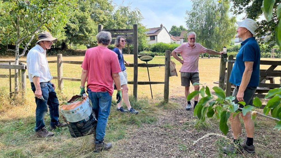 A group of five people stood chatting in a circle in a field, there is a rake balanced up against a fence that runs through the centre of the picture and a man holding a bag of garden waste.