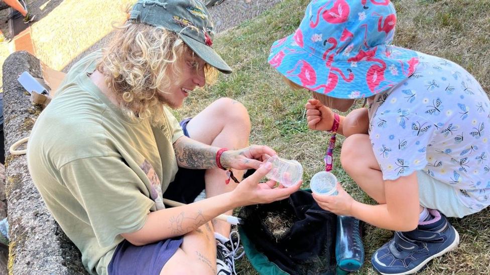 A person showing a child a carton with a bug in it. Both are sat in the grass. The man is wearing a green shirt and hat and the child is wearing a blue bucket hat with flamingoes.