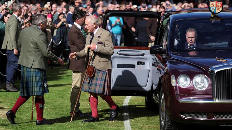 King Charles wearing a kilt, exits a Bentley which is parked on a stretch of grass. He is being greeted by an official and crowds look on in the background