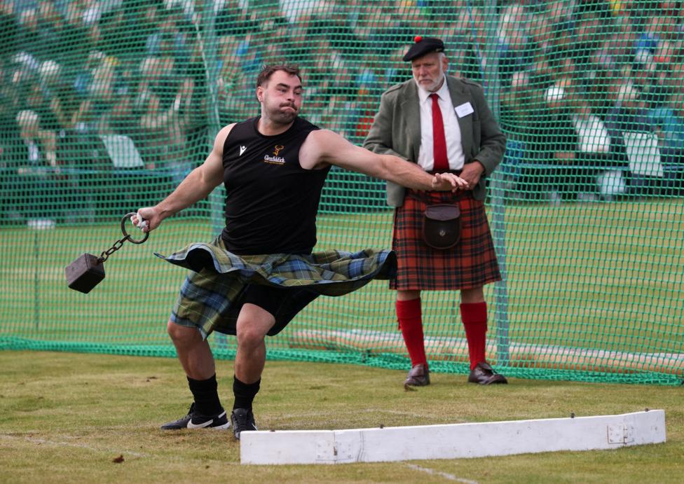 a man in a kilt and sleeveless top throws a heavy weight while a kilted judge and spectators watch