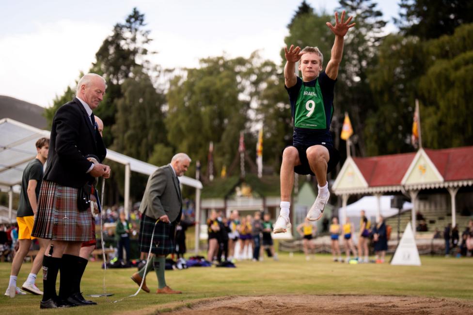 a man in a kilt watches on a boy long jumps into a sand pit