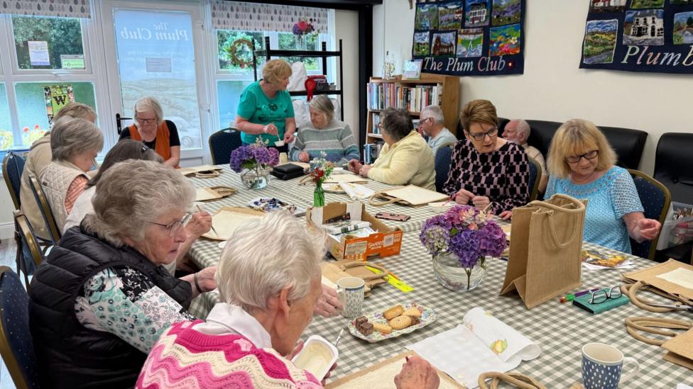 A group of older adults gathered round a large table at the Plum Club meeting place. They are completing a craft activity using paint, napkins and bags.