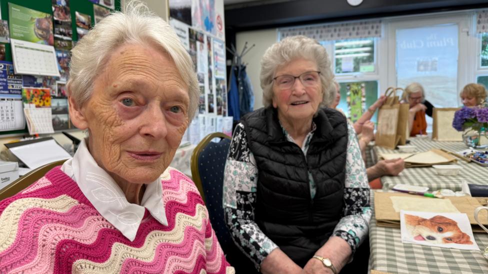 Two older women smiling at camera. The woman in the foreground wears a bright pink patterned jumper and white shirt, while the woman beside her wears glasses, a black padded vest, and a floral top. Behind them, people sit around tables covered with craft supplies, fabrics, and other activities.