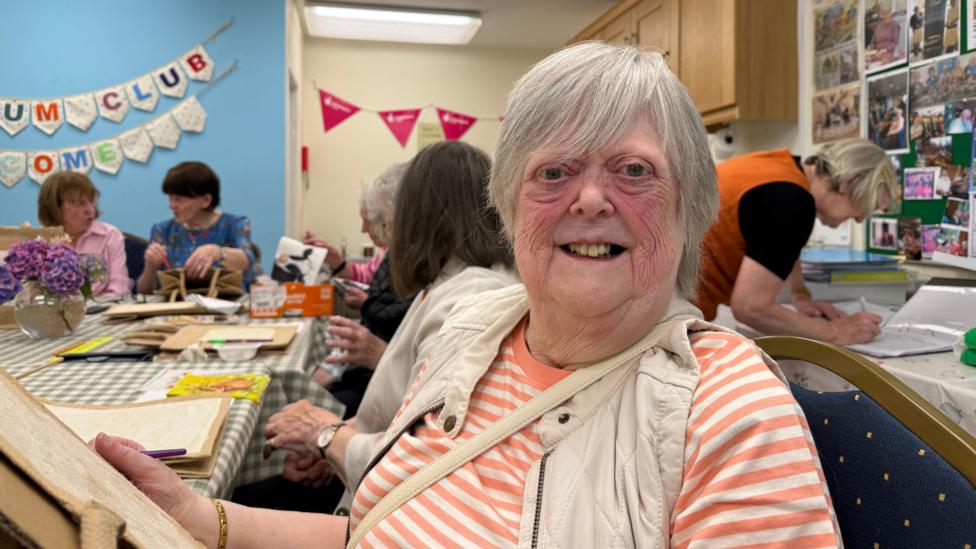 Hilary smiles at the camera while seated. She is wearing a striped orange-and-white top with a light quilted vest. Other people are working away in the background on a craft activity. She is holding a bag and Anne Bradley is in the background wearing a orange dress.