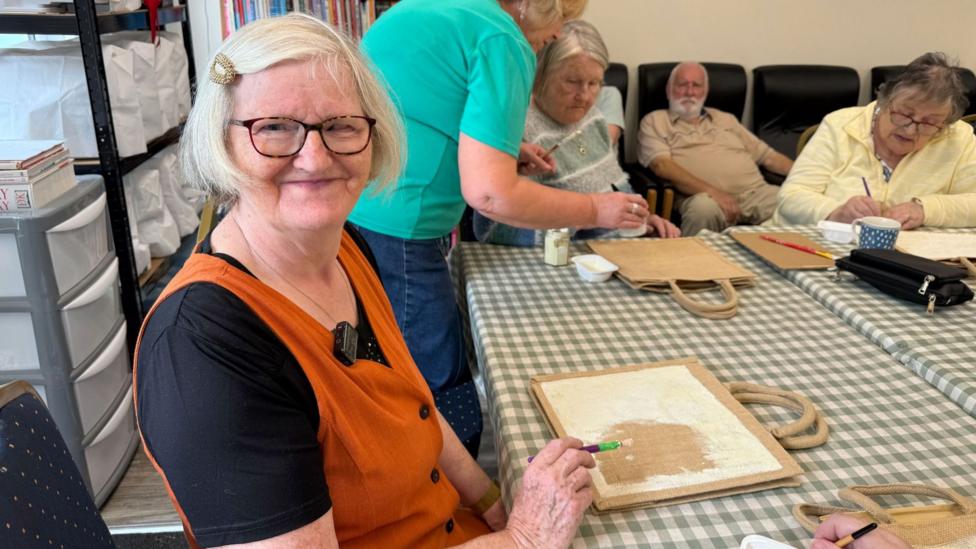 A group of older adults takes part in a craft activity around a checkered table in a community room. Anne is the woman smiling at the camera wearing an orange dress. She has a black shirt below it.