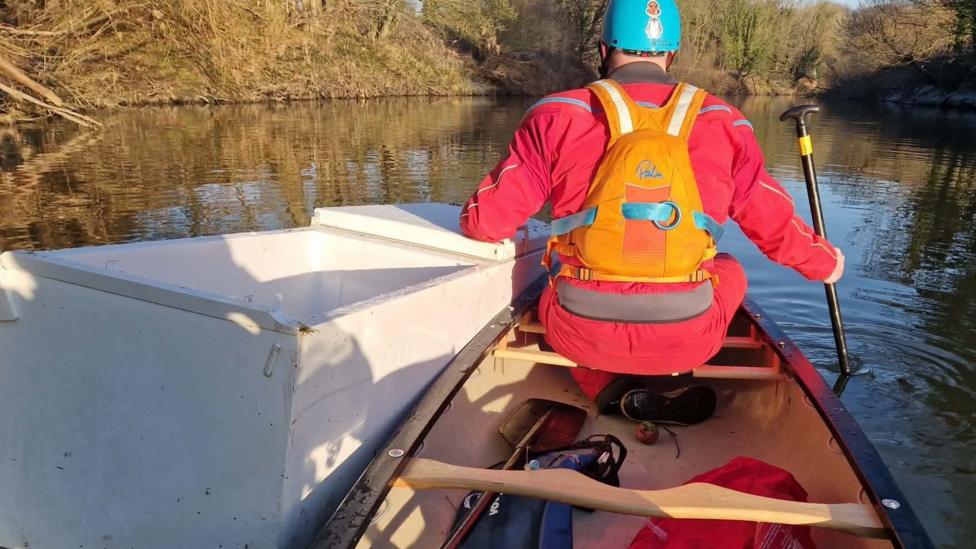 A man is holding on to a fridge while paddling a canoe along the River Wear. The fridge is alongside the canoe and the man is wearing an orange life-jacket over a red coat and a blue helmet.