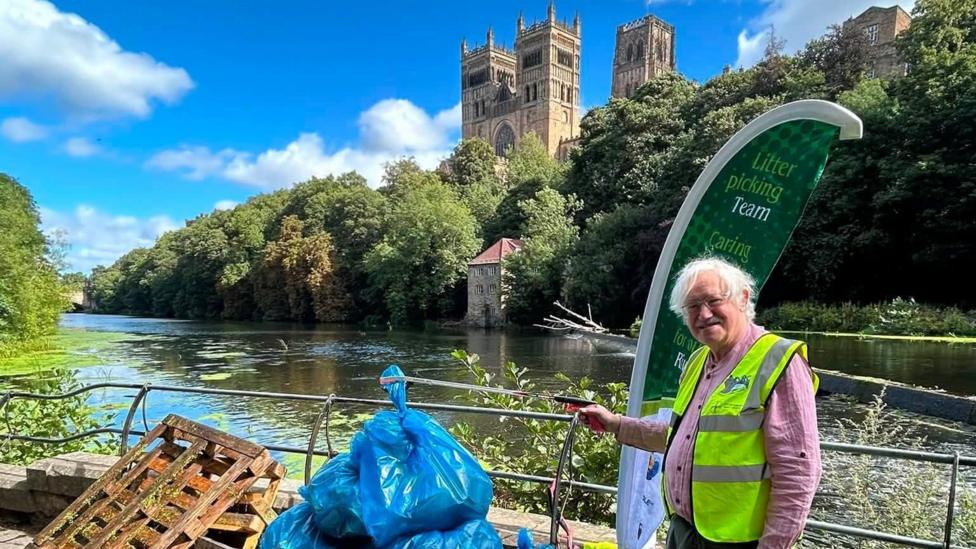 A litter picker in hi-vis is stood on the banks of the River Wear with Durham Cathedral on the hill behind him. He is smiling and depositing a blue bag of rubbish on a pile near the river railings.