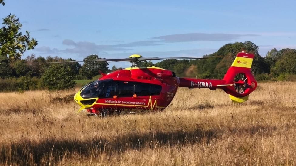 A midlands air ambulance which is red and has a yellow nose, tail and roof. it is parked in a field of brown grass