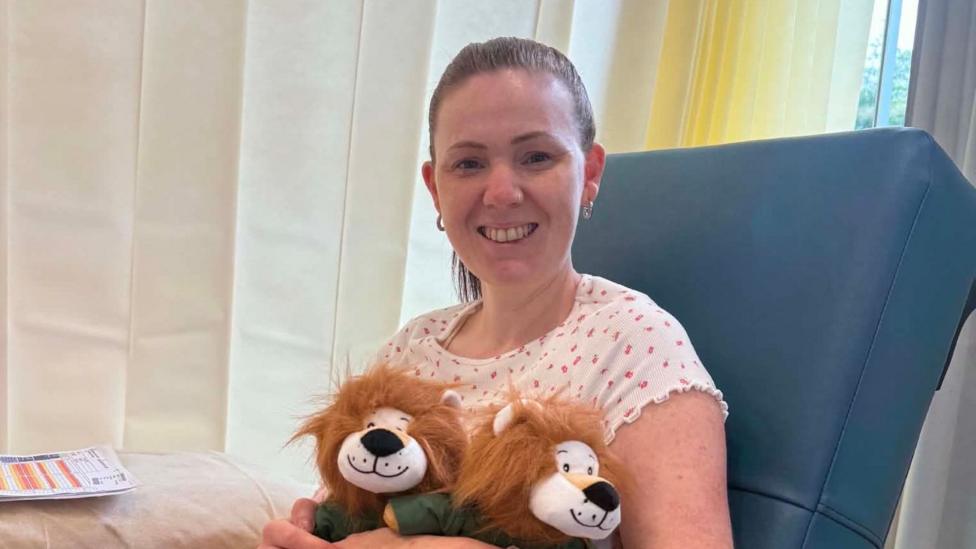 Rhiannon is wearing pajamas and sitting in a chair in hospital holding two cuddly toys.