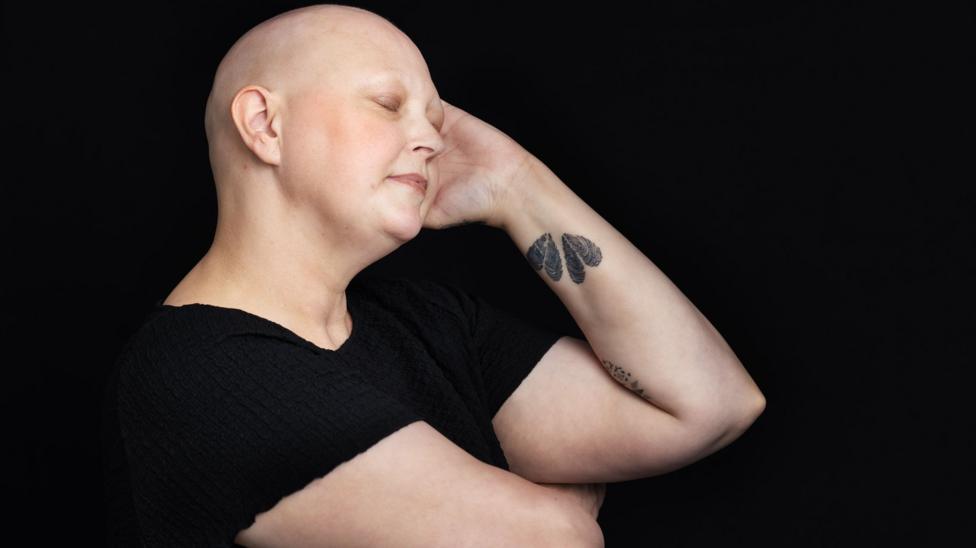 Sam is in profile and running her hand over her bald head. She is wearing a black T-shirt and has a tattoo on her forearm.