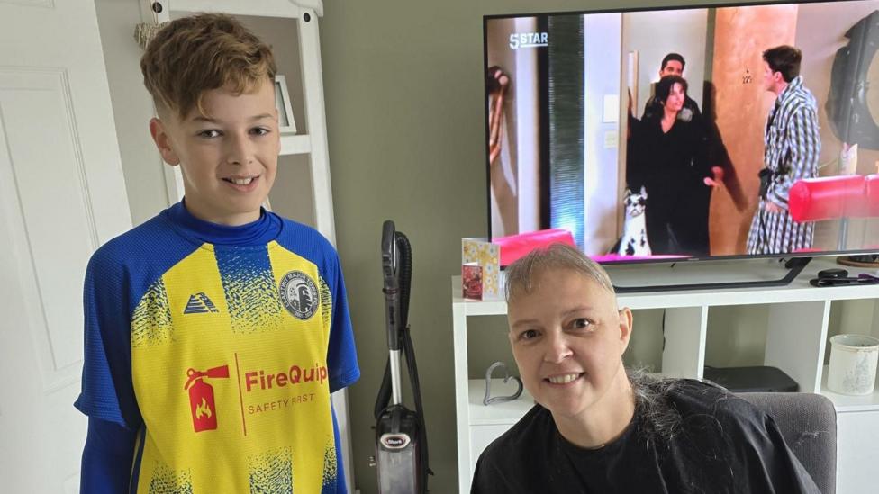 Sam and her son after having her head shaved. Her young son is wearing a football kit. She has just hair at the front of her head remaining and is wearing a black hairdressing cape.