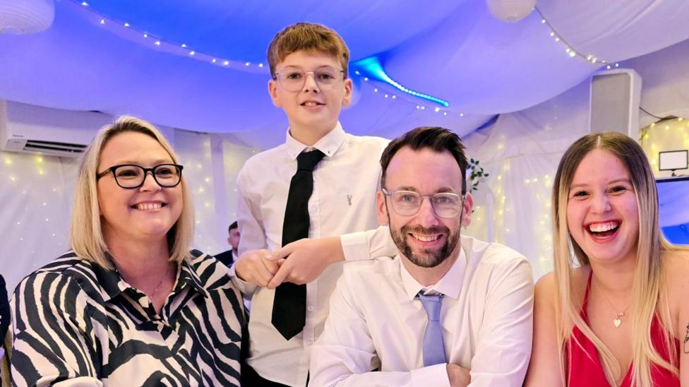 Sam at an event with her husband and children. They are all smiling. She is wearing a black and white print top and has a blonde bob.Her son and husband are wearing white shirts and ties. Her daughter has long blonde hair and is wearing a red top.