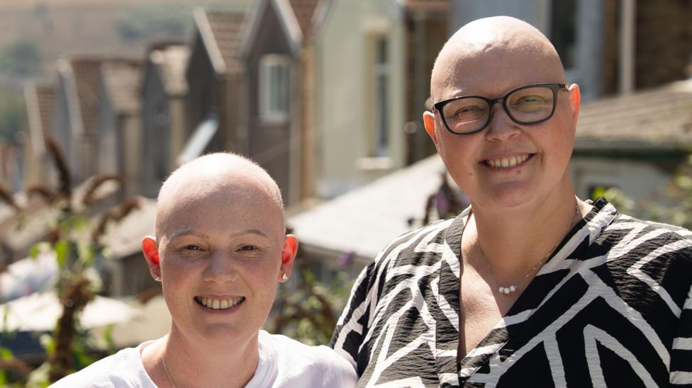 Rhiannon and Sam standing on a residential street, smiling at the camera. Both have bald heads. Rhiannon is wearing a white T-shirt and Sam is wearing a black and white print dress.