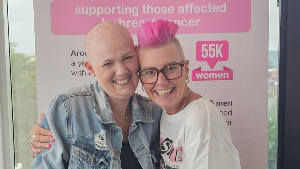 Lisa has her arm around Rhiannon and both are looking at the camera and smiling. Lisa has spiky bright pink hair and her head shaved at the sides. She is wearing a white T-shirt. Rhiannon is bald and wearing a denim jacket.