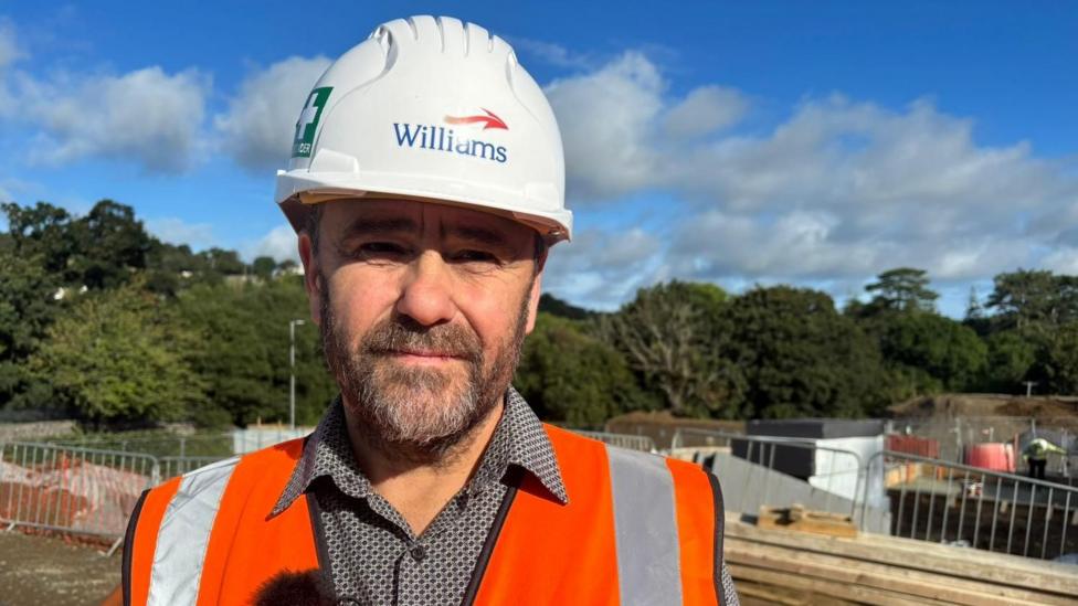 A picture of a man wearing a white hard hat and a bright orange high vis jacket. He's looking at the camera and standing on a building site. There are fences and trees behind him