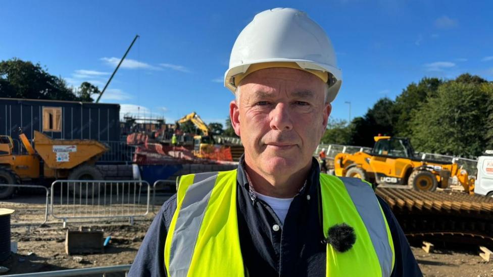 A picture of a man wearing a white hard hat and a bright yellow high vis jacket. He's looking at the camera and standing on a building site. There are fences and heavy machinery behind him including a dump truck and a crane.