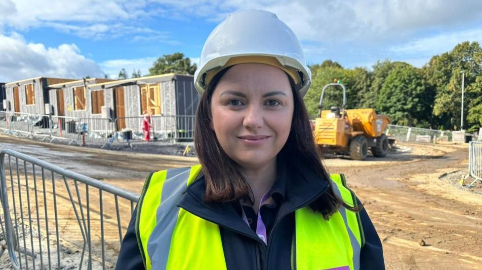 A picture of a woman wearing a white hard hat and a yellow high vis jacket. She's looking at the camera and standing on a building site. There's a dumper truck behind her and also homes that are being built.