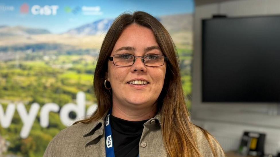 A picture of a woman wearing a light brown coloured shirt and a black tshirt. She's wearing glasses and a lanyard around her neck. Behind her is a TV that is turned off and a large sign showing outside and a lot of greenery. She is smiling at the camera.