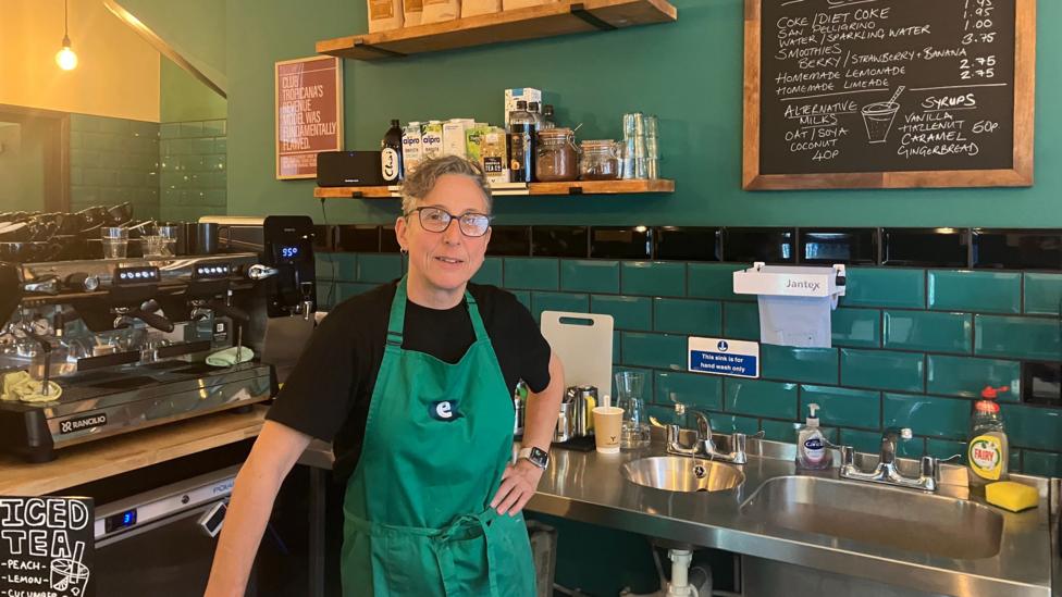 A woman wearing a dark shirt and green apron stands behind the counter of a café, next to a coffee machine and board advertising drinks