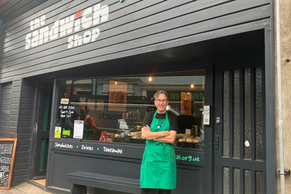 A woman wearing a green apron and dark t-shirt stands outside a café, which has sandwiches for sale in the front window