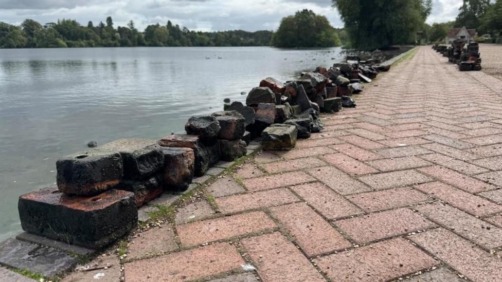 There is a lot of broken bricks laid up in a line next to a lake. They are on top of a black brick outline and a red brick inside.