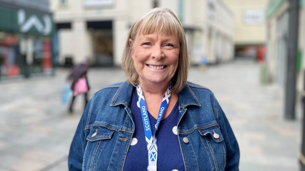 Sharon has a blonde bob and a fringe. She is smiling, wearing a blue t-shirt with a denim jacket, and is standing in a high street.