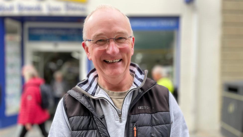 Andrew has short receding grey hair and wears rimless glasses. He is wearing a grey quarter-zip jumper and a black gilet, and is standing in front of a shop with people passing by on a high street.