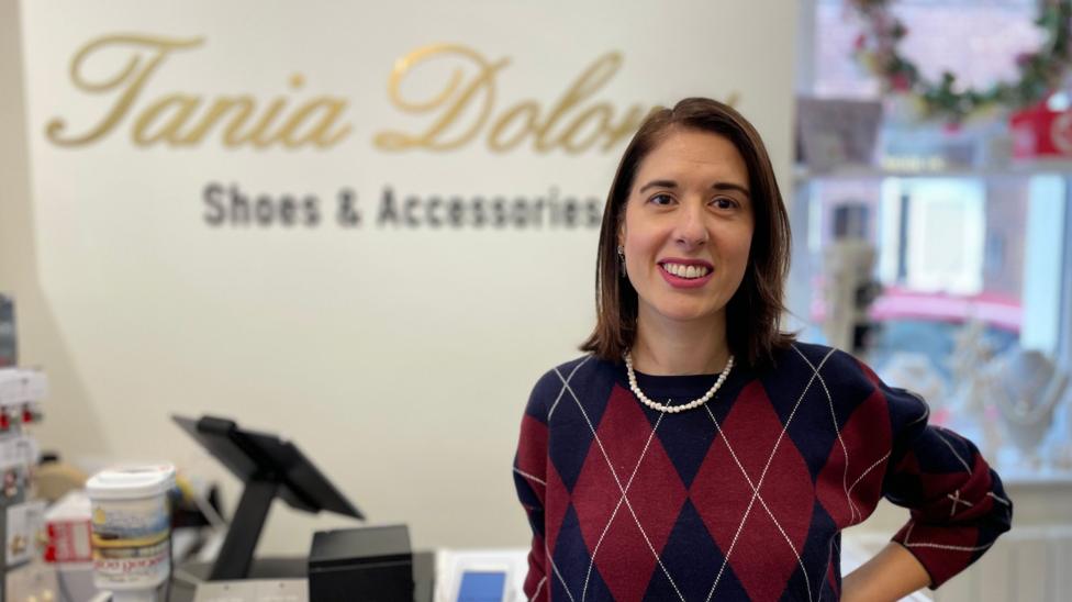 Tania Dolores has shoulder-length brown hair and is smiling. She is wearing a navy and red diamond-patterned jumper. She is standing in front of a till with a gold sign of her name on the wall behind her.