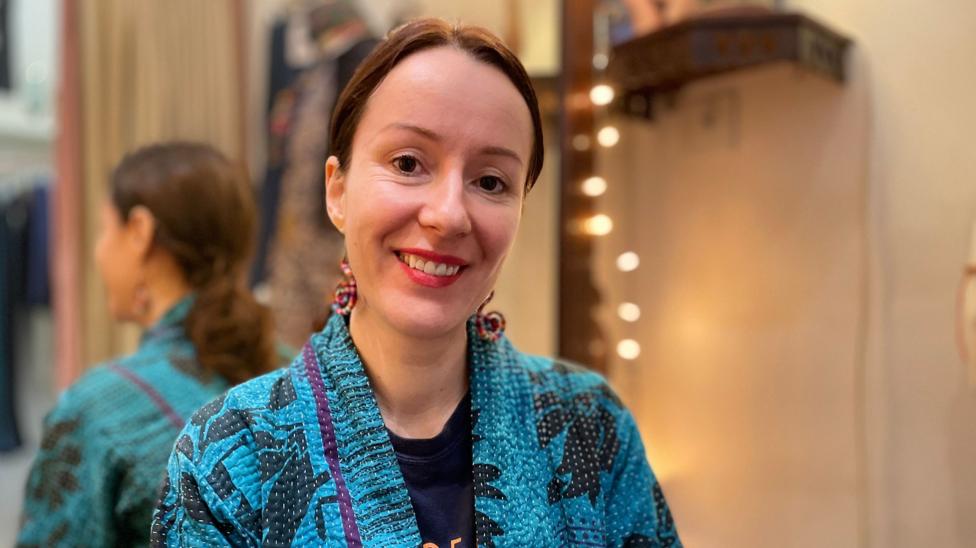 Linda Murphy has brown hair tied back and is wearing a blue and black patterned robe. She is smiling and sitting in front of a mirror on the left with fairy lights to the right.